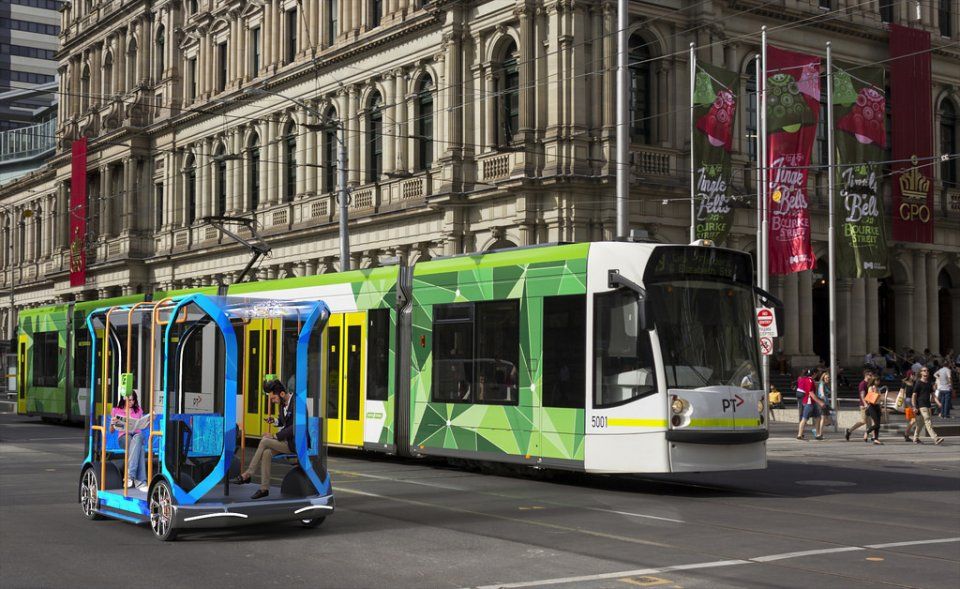 Modern public transportation scene featuring a green tram alongside a small, futuristic, autonomous vehicle on a bustling urban street with a historic building in the background.